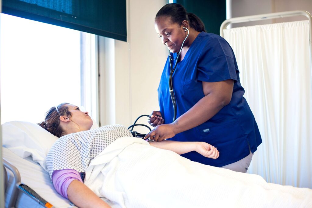 A woman is standing taking the blood pressure of another woman who is a patient in a hospital bed.
