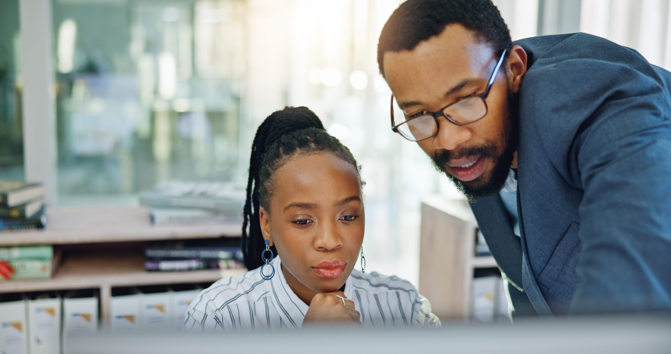 Two people working together at a desk in an office setting, looking at a computer screen.