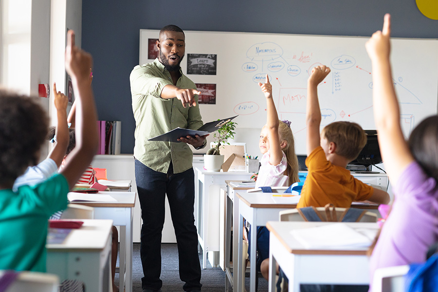 Black male teacher standing in front of a classroom with children raising hands